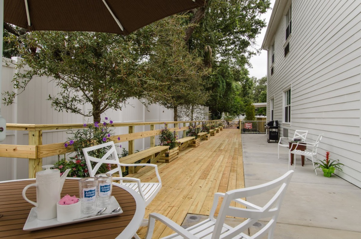 A peaceful outdoor patio with white chairs and tables, set against a wooden deck and surrounded by trees.