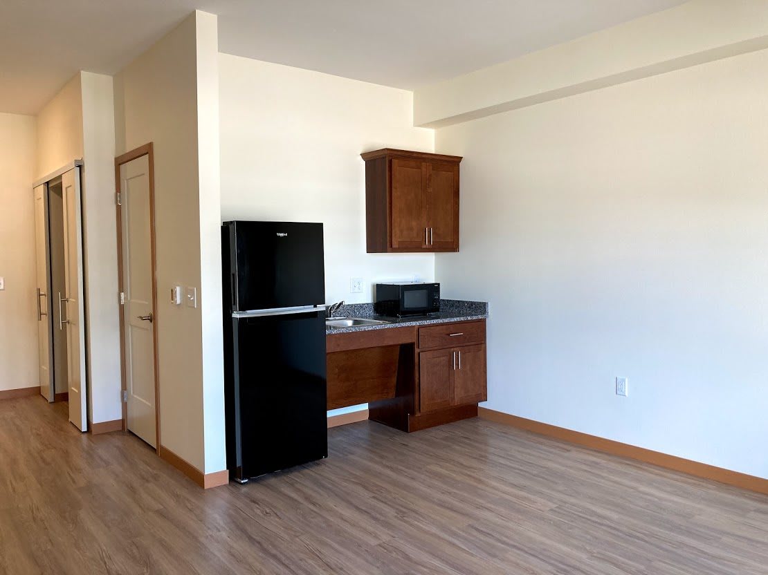 A minimalist apartment kitchen with a refrigerator, microwave, and wooden cabinetry.