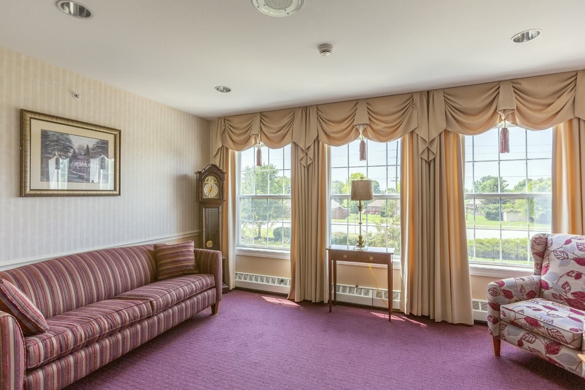 A traditional living room with a striped sofa and grandfather clock, featuring large windows with drapes.
