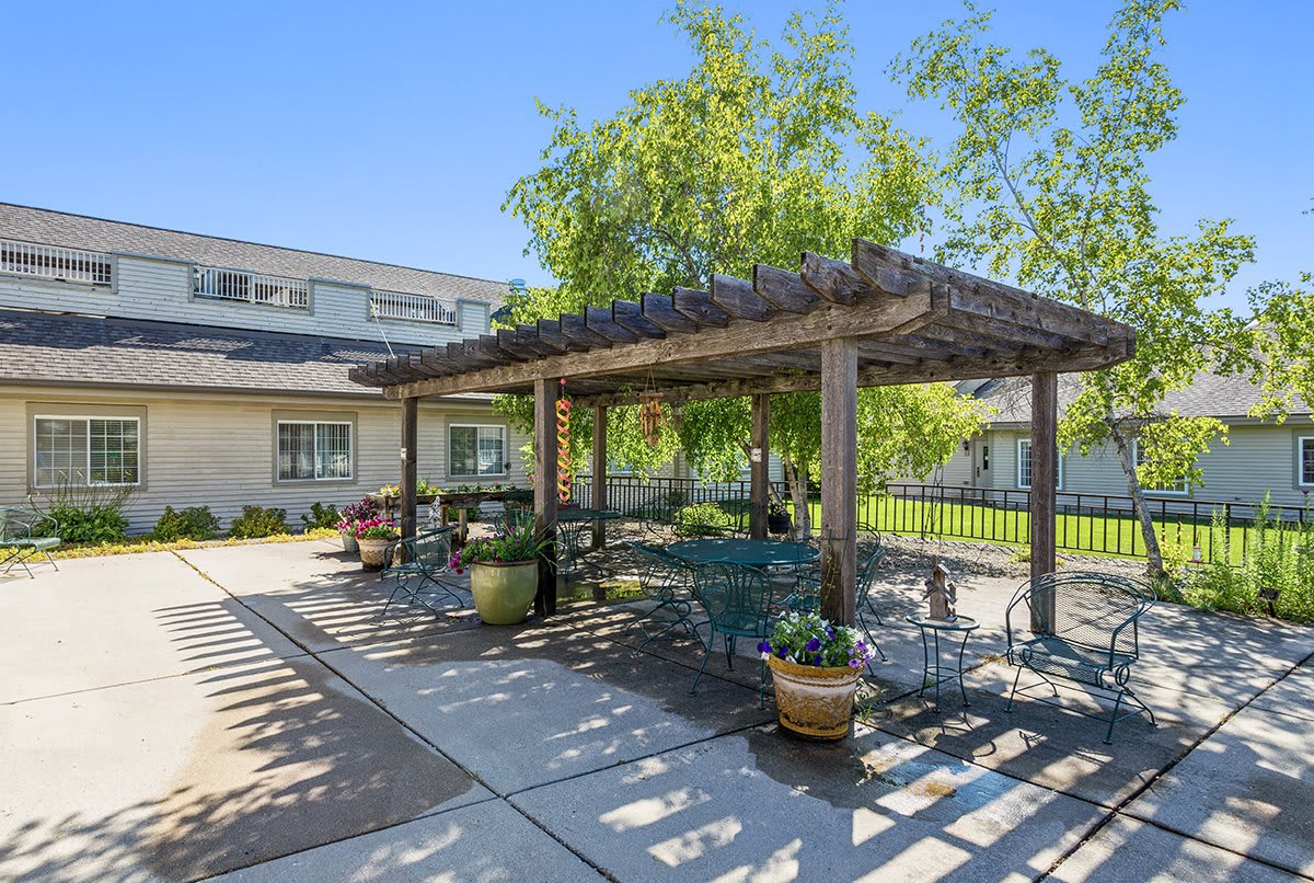 A sunny outdoor patio area with a pergola and potted plants, providing shaded seating next to a building.