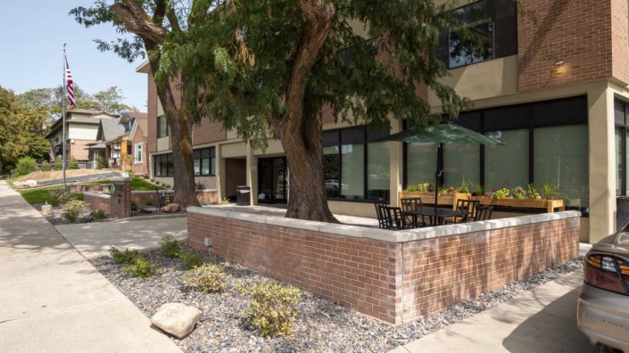 An outdoor patio with tables and chairs, surrounded by a brick wall and trees along a sidewalk.