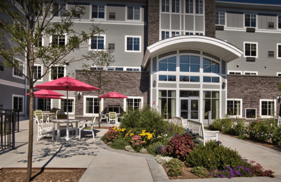 A building with outdoor seating and umbrellas, surrounded by landscaped gardens and flowers near the entrance.