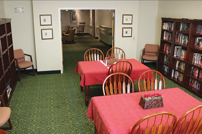 A cozy indoor dining area with tables covered in red tablecloths, wooden chairs, and a bookshelf against a carpeted floor.