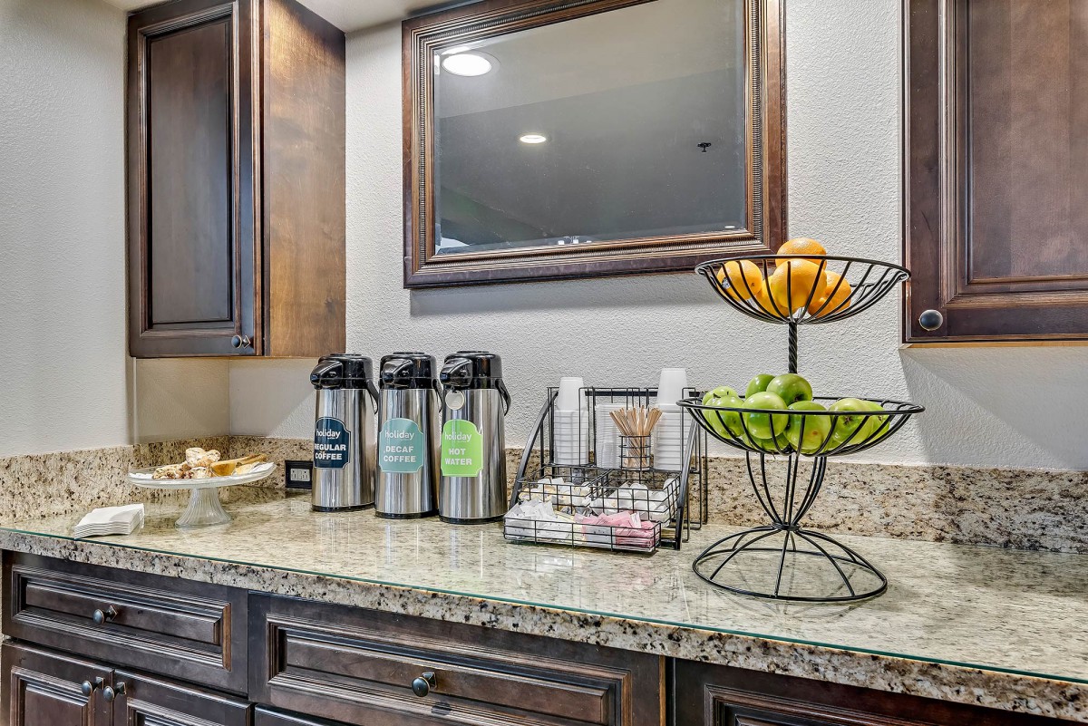 A well-organized coffee station with beverage dispensers, a fruit basket, and cups on a countertop.