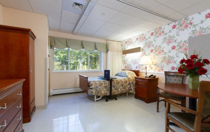 A cozy hospital room with a single bed, wooden furniture, and a floral wallpaper design.