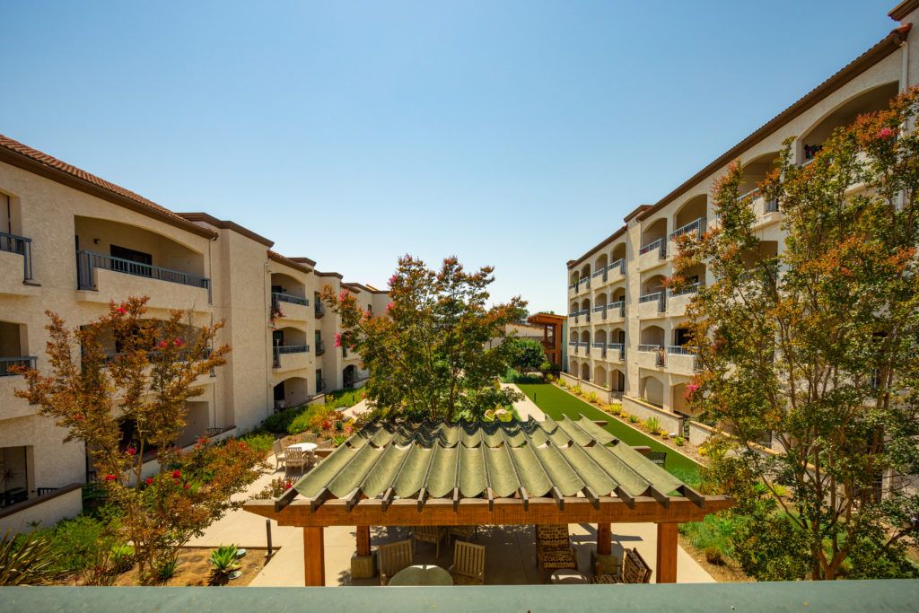 A view of apartment buildings surrounding a landscaped courtyard with an outdoor patio on a sunny day.