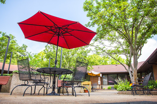 A bright red umbrella provides shade over a patio table and chairs, surrounded by greenery.