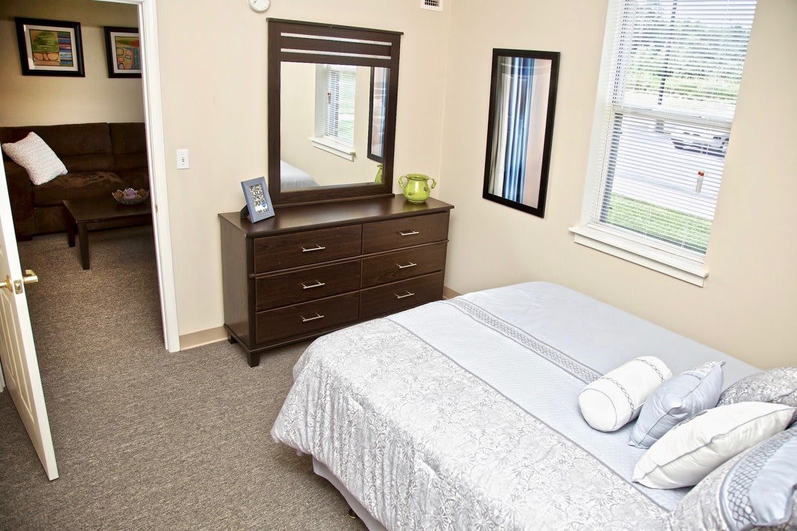 A cozy bedroom with a neatly made bed, a wooden dresser with a mirror, and natural light coming through the window.