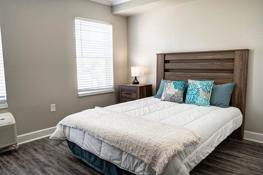 A cozy bedroom with a neatly made bed, pillows, and a nightstand with a lamp beside a window.
