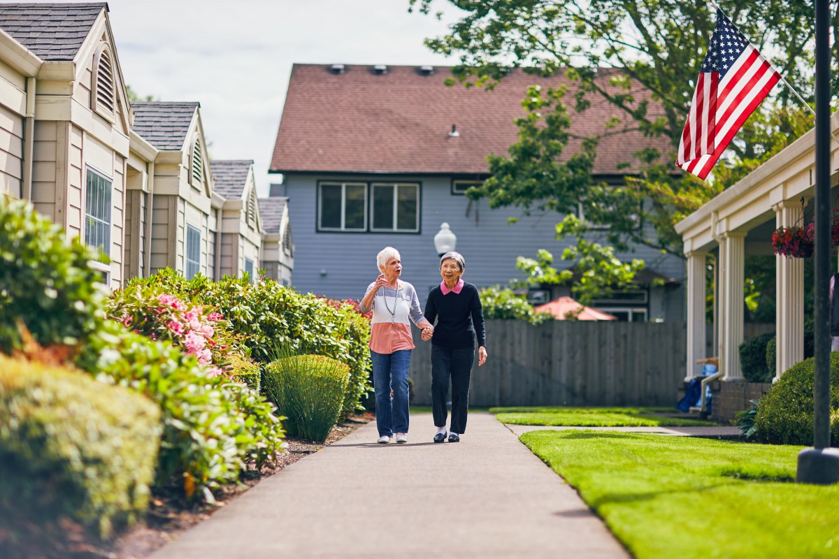 Two elderly people walking together along a garden pathway in a community setting on a sunny day.