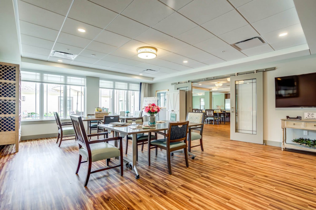 A modern dining room with wooden flooring, featuring a set of tables and chairs, illuminated by ceiling lights and surrounded by large windows.