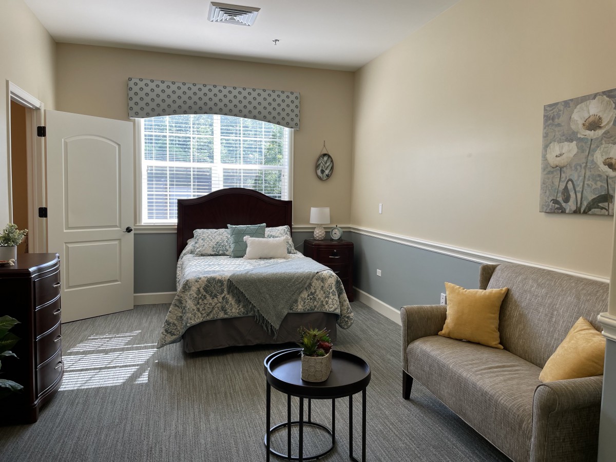 A cozy bedroom with a bed, sofa, and natural lighting from a large window.