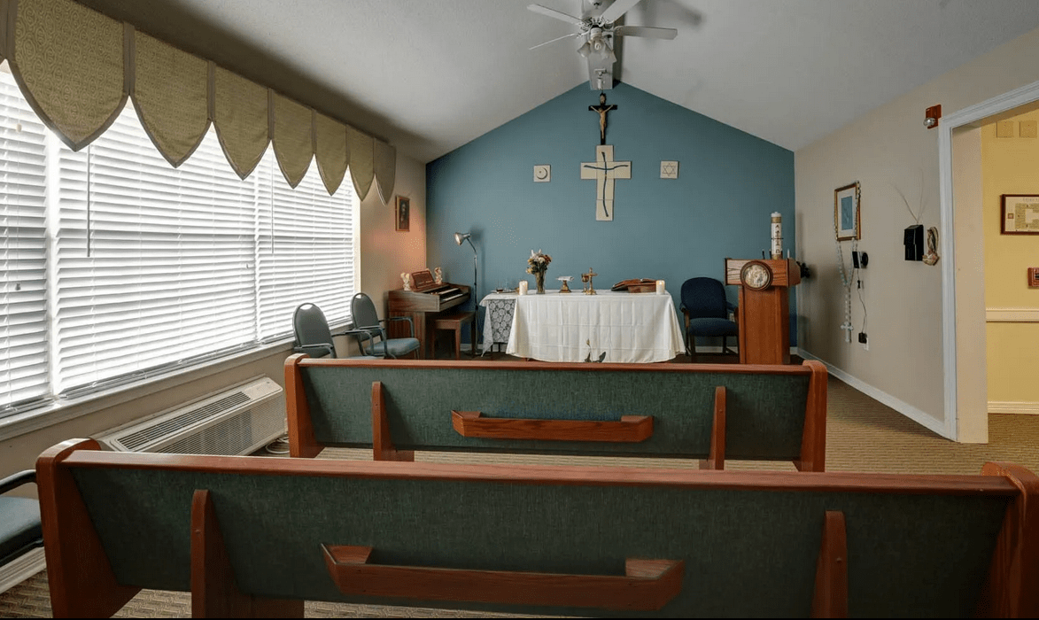 A small chapel interior with church pews, a cross on the wall, and an altar at the front.