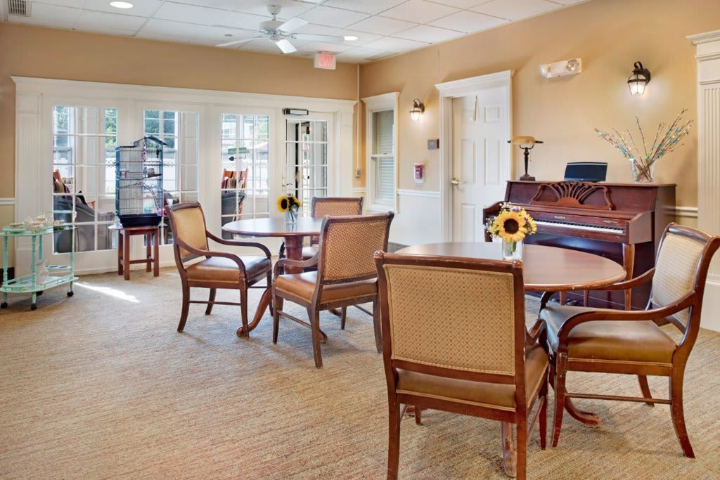 A spacious room with chairs and tables arranged around a grand piano, featuring large windows and soft lighting.