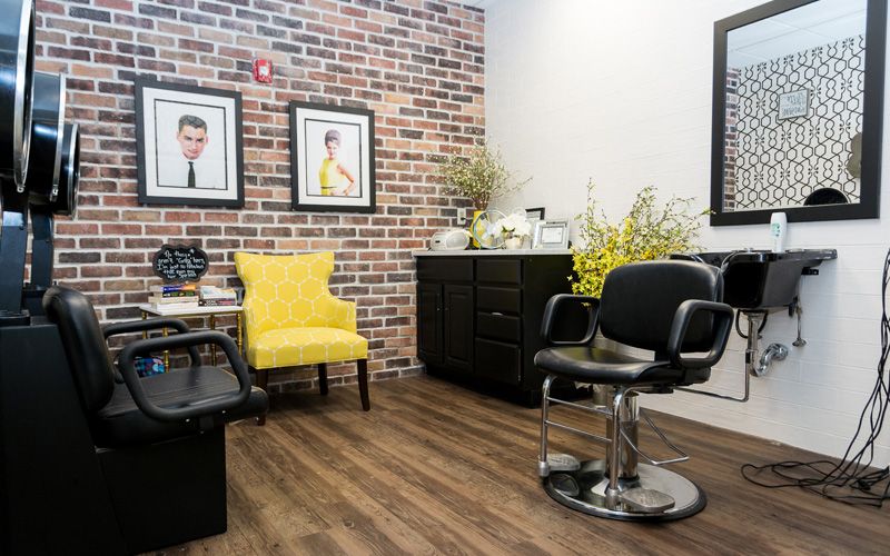 A modern hair salon interior with a yellow armchair and black styling chair, featuring a brick accent wall and decorative elements.