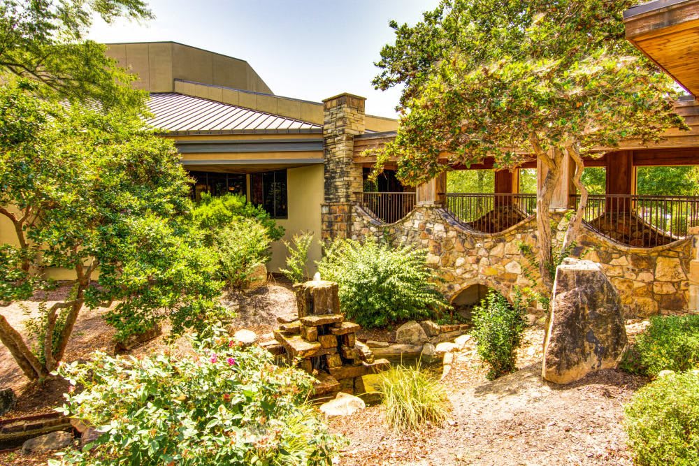 A tranquil garden landscape featuring lush greenery, a stone building, and a small water feature.