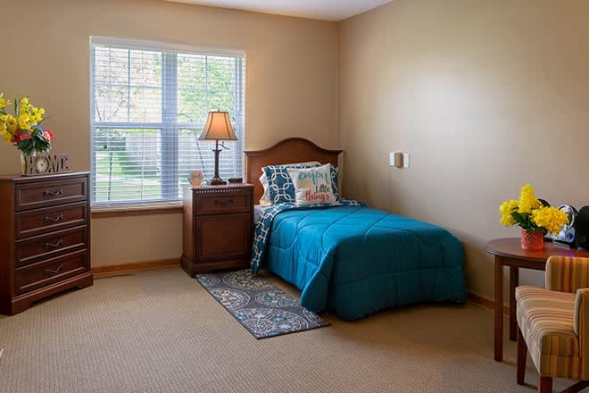 A cozy bedroom with a single bed adorned in teal bedding, a wooden headboard, and a nightstand with a lamp beside a large window.
