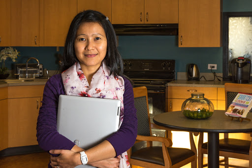 A woman holding a laptop, smiling in a cozy kitchen setting.