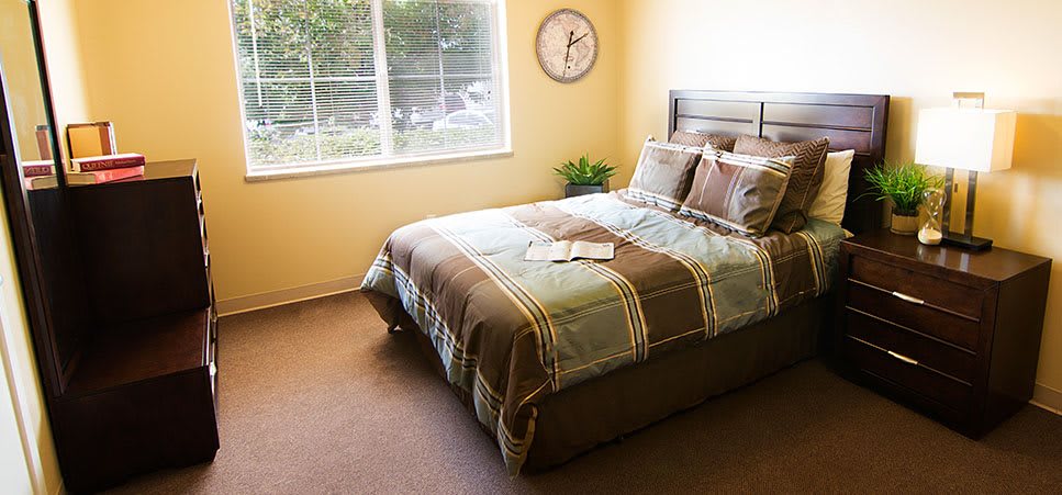 A cozy bedroom with a neatly made bed, a wall clock, and a window letting in natural light.