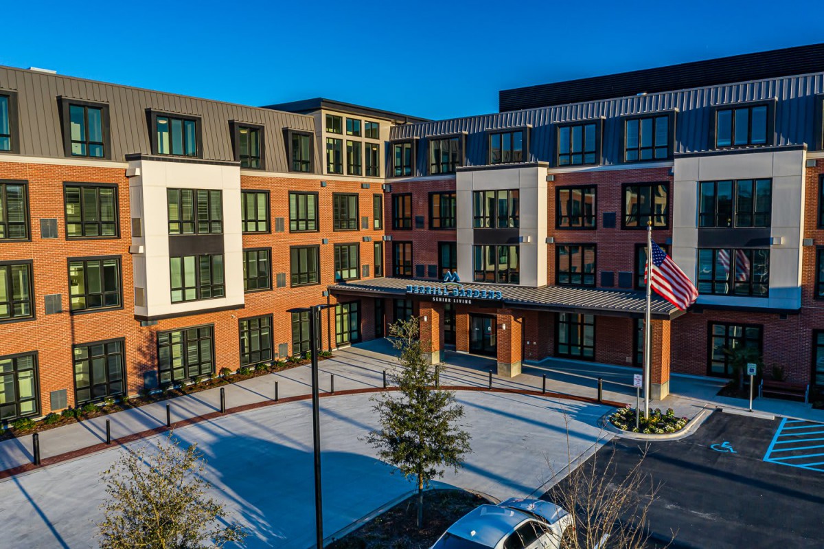A modern apartment complex with large windows and an entrance area, featuring a flagpole and landscaped surroundings.