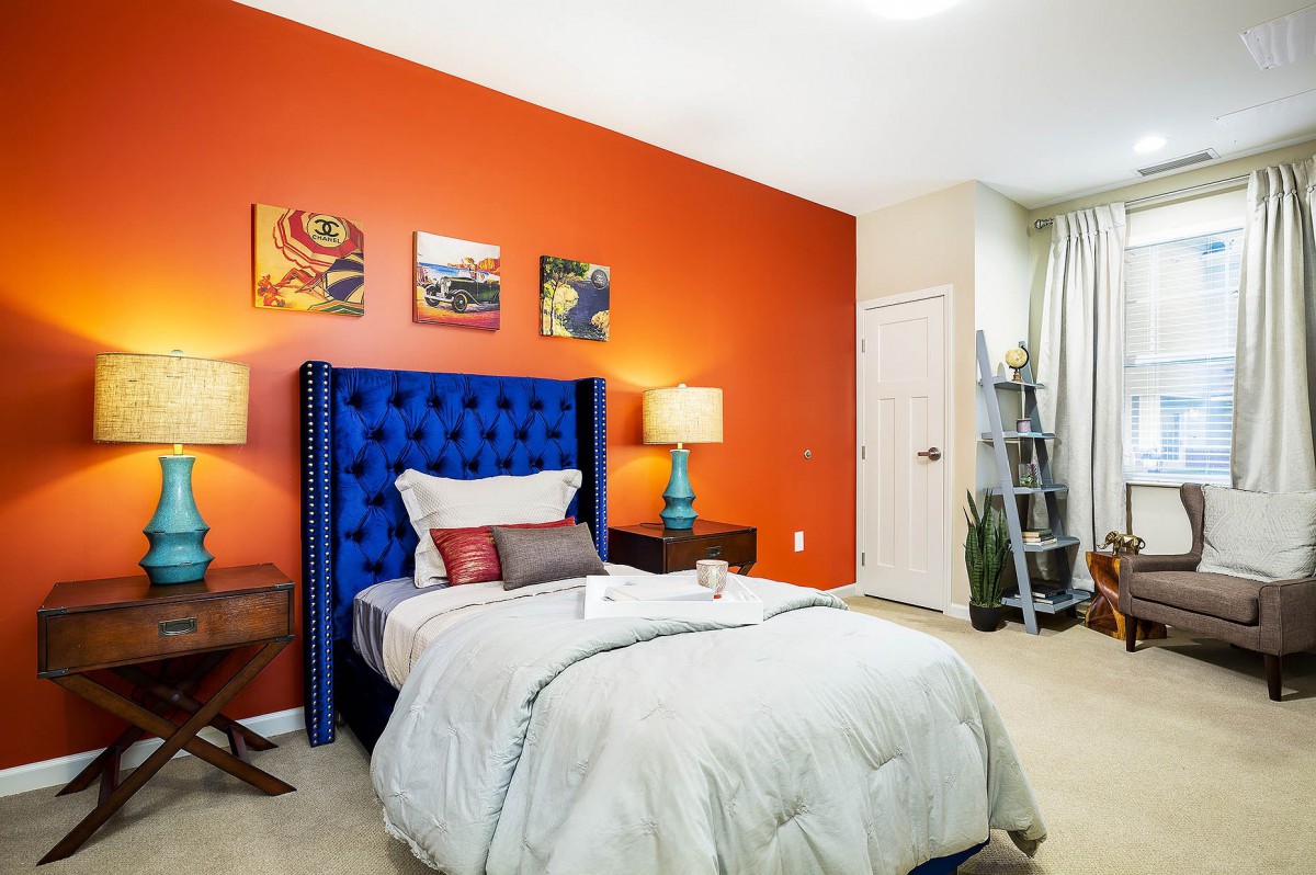 A modern bedroom with a striking orange accent wall and a blue upholstered headboard, featuring stylish decor and warm lighting.