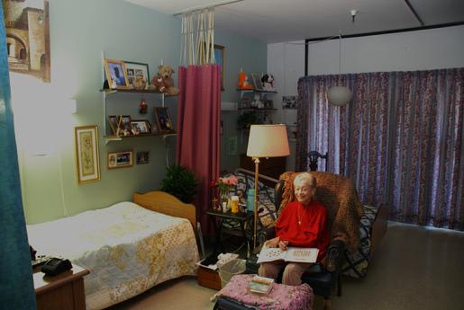 An elderly woman sitting in a cozy room decorated with various personal items and furniture.