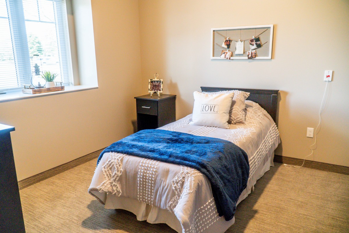 A cozy bedroom with a single bed, blue blanket, and decorative pillows next to a window and nightstand.