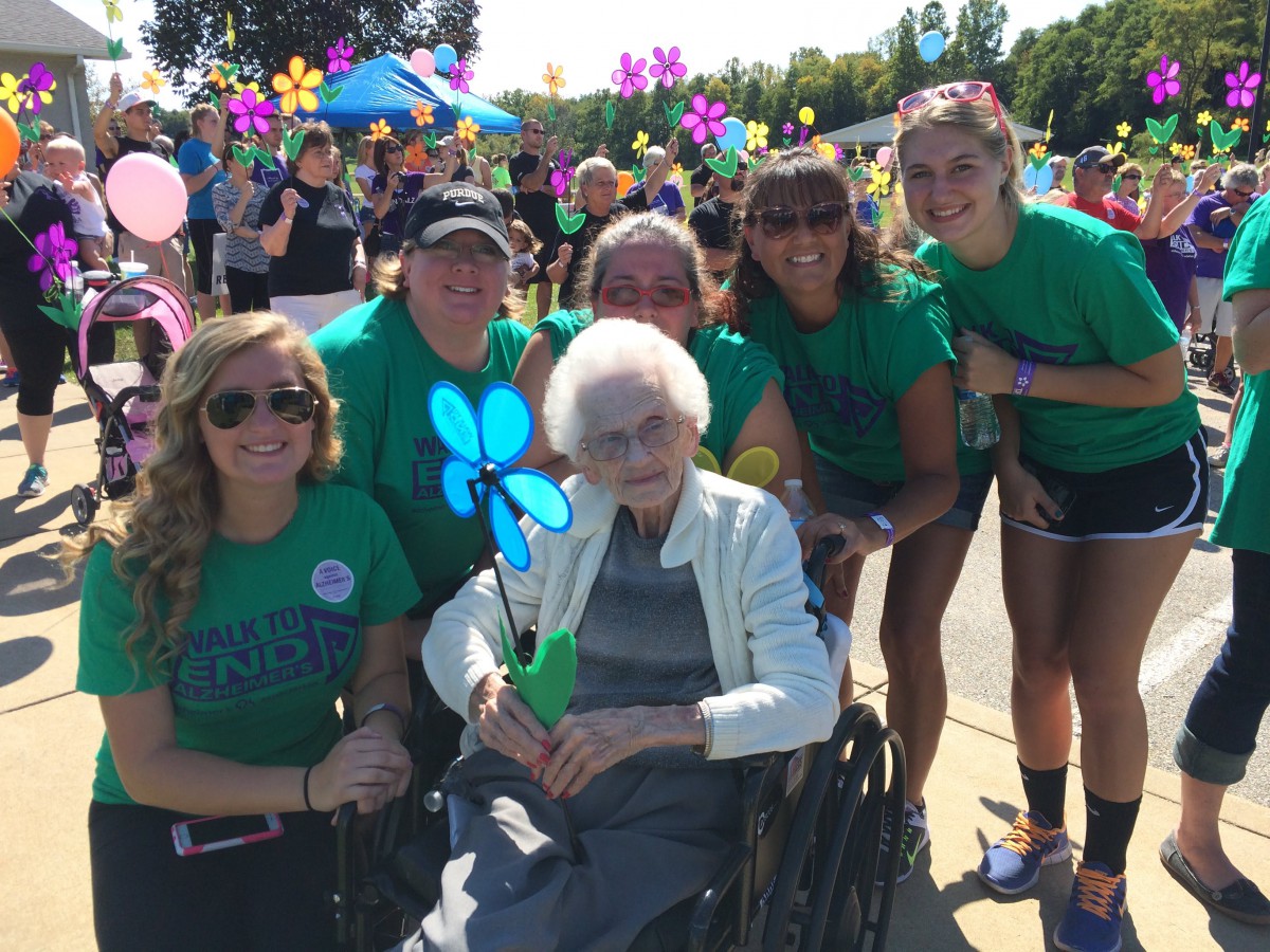A group of people gather around an elderly woman in a wheelchair during a colorful outdoor event.