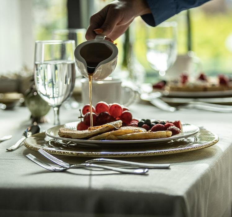 A hand pouring syrup over pancakes topped with fresh berries on a elegantly set table.