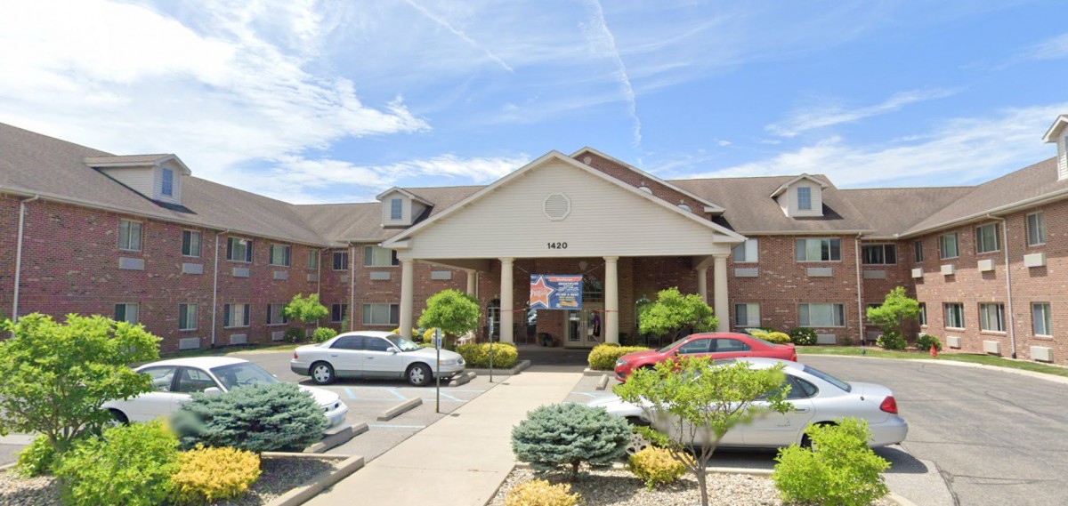 A view of a building entrance with parked cars and landscaped greenery under a partly cloudy sky.
