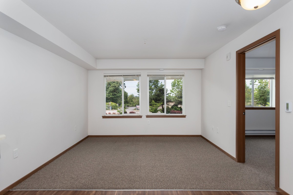 An empty room with carpeted flooring and two large windows overlooking greenery.