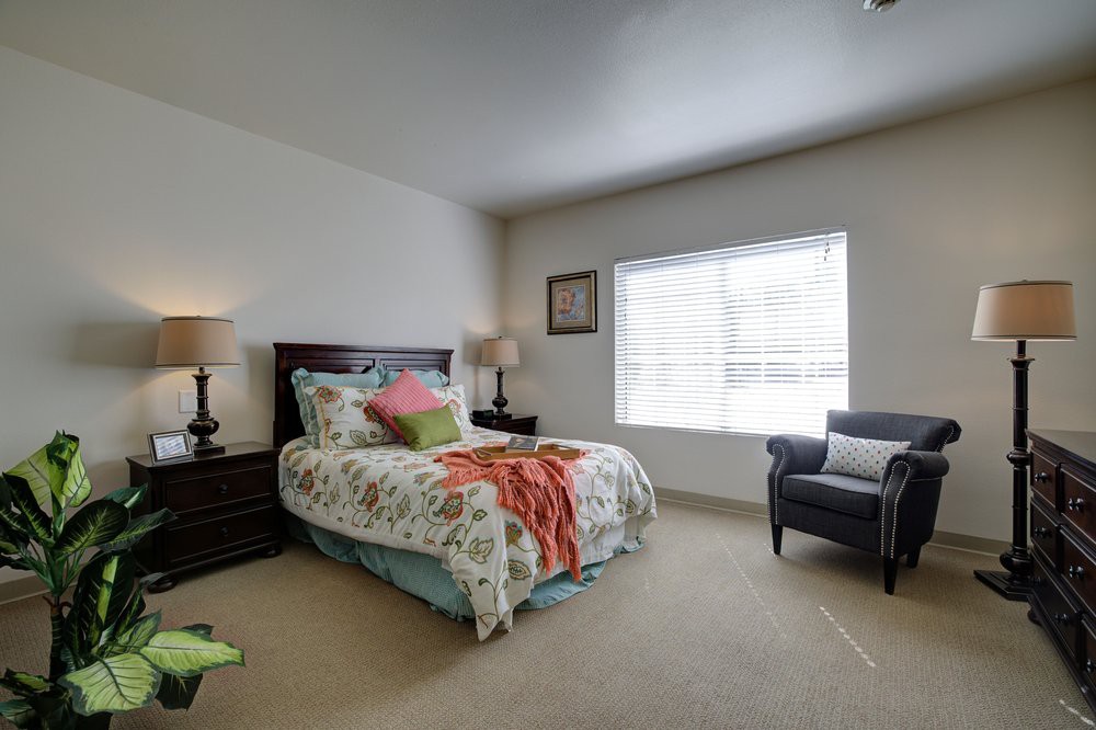 A cozy bedroom with a neatly made bed, bedside tables with lamps, a comfortable chair, and a large window letting in natural light.