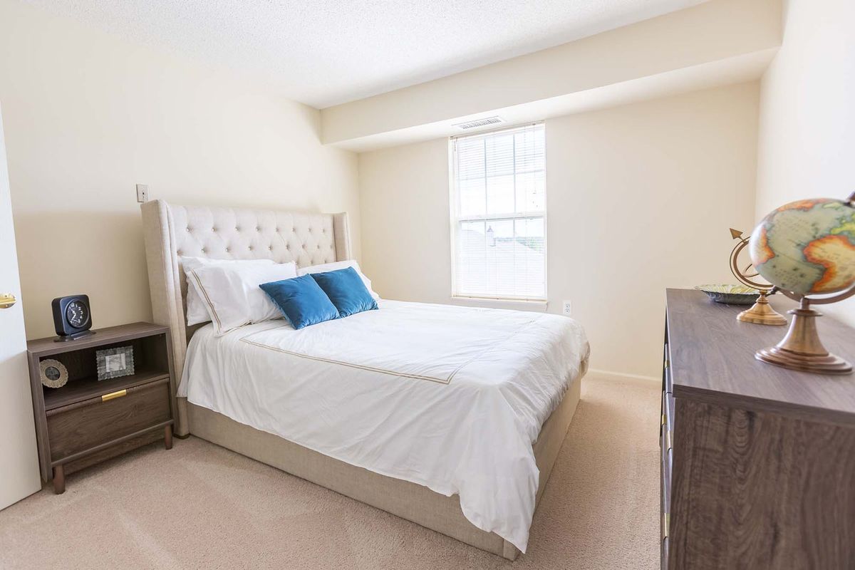 A bright and simple bedroom with a neatly made bed, two blue pillows, and a window bringing in natural light.