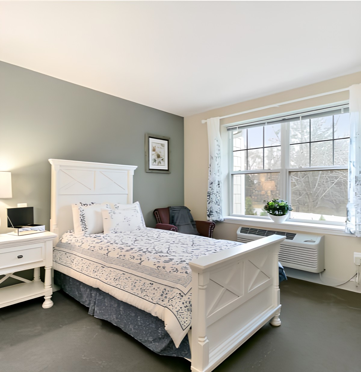 A cozy bedroom with a large window, featuring a bed, nightstand with a lamp, and simple decor.