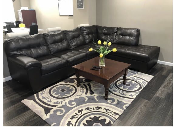 A cozy living room featuring a black leather sectional sofa, a wooden coffee table with yellow tulips in a vase, and a patterned rug.