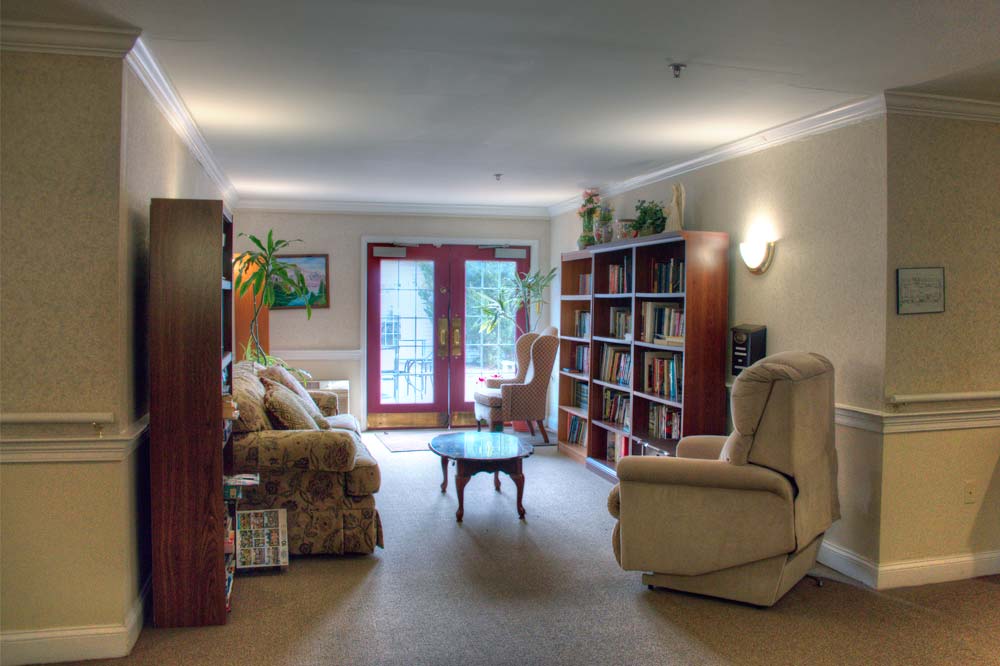 A cozy living room with armchairs, bookshelves, and a coffee table, illuminated by natural light from large glass doors.