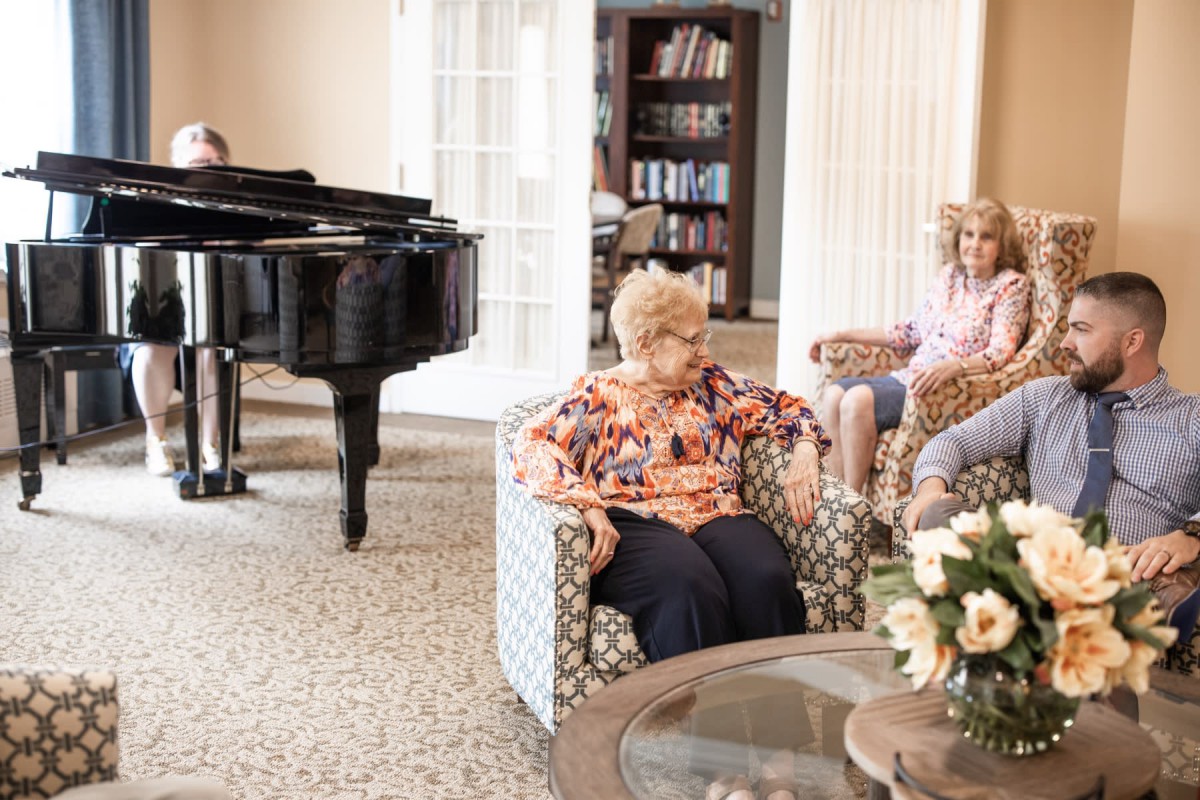 A cozy living room with seniors relaxing in armchairs, alongside a grand piano and bookshelves.