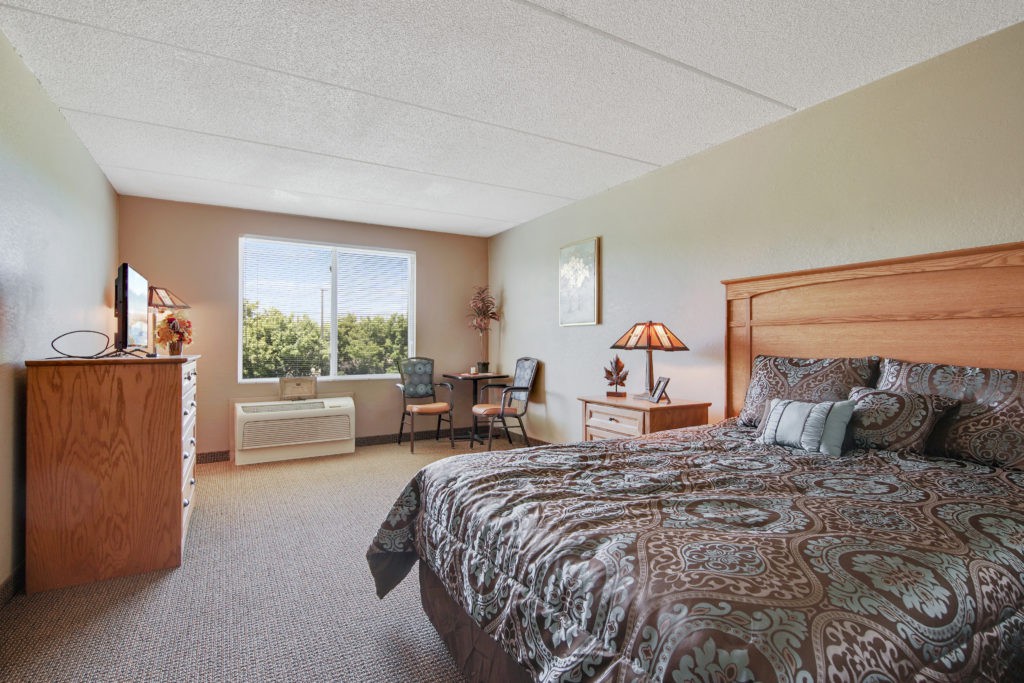 A well-lit bedroom featuring a comfortable bed, a desk with a lamp, and a large window offering natural light.