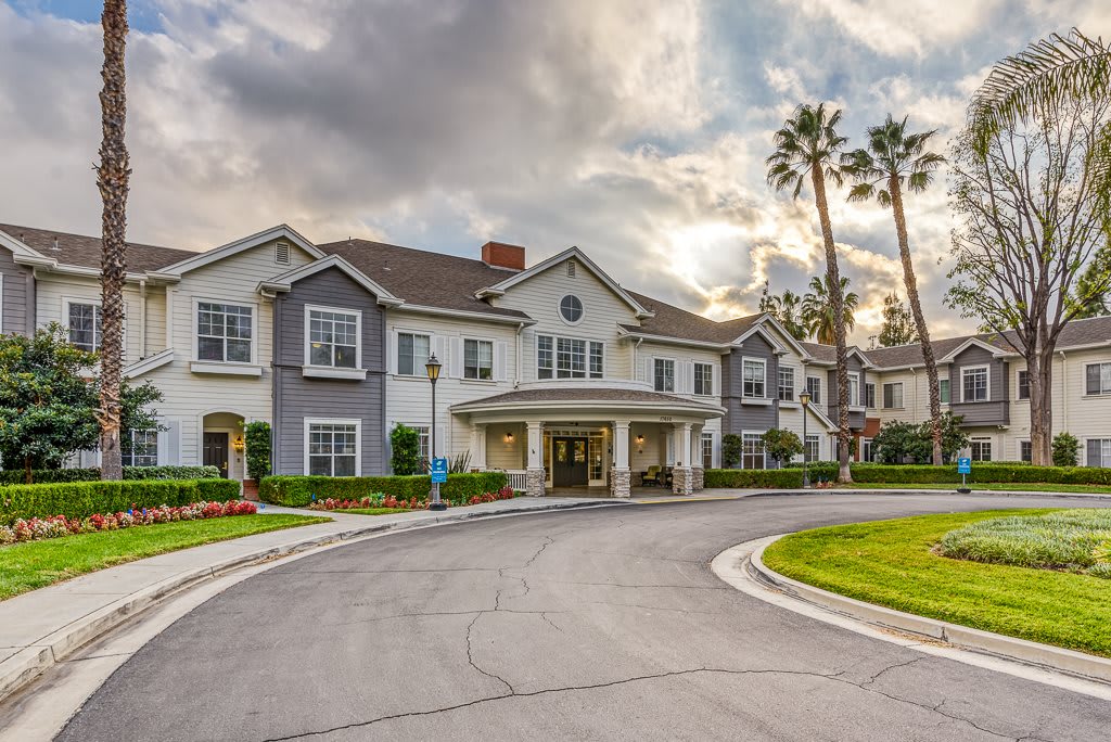 A large residential building with a circular driveway, surrounded by lush landscaping and tall palm trees under a cloudy sky.