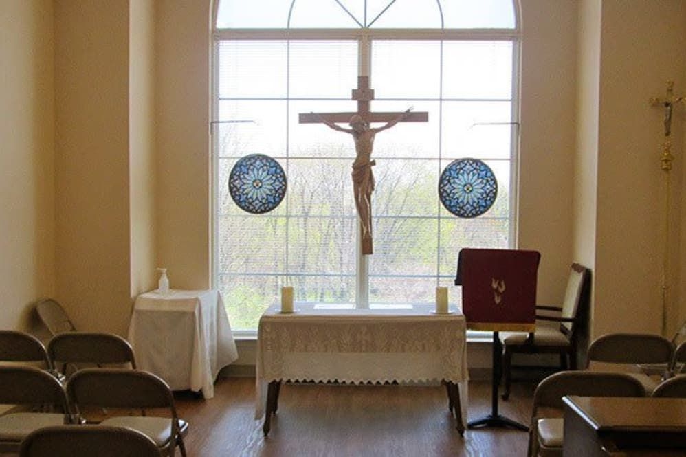 A small chapel featuring an altar with a crucifix, flanked by chairs and illuminated by natural light through a large window.
