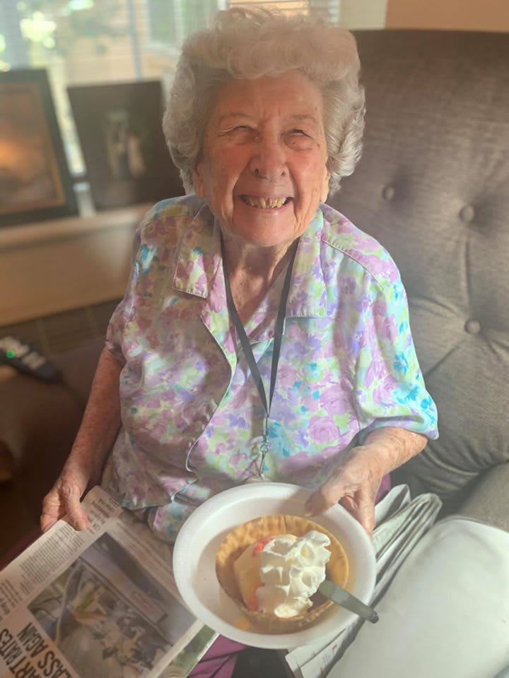An elderly woman sitting and smiling while holding a plate with a pie.
