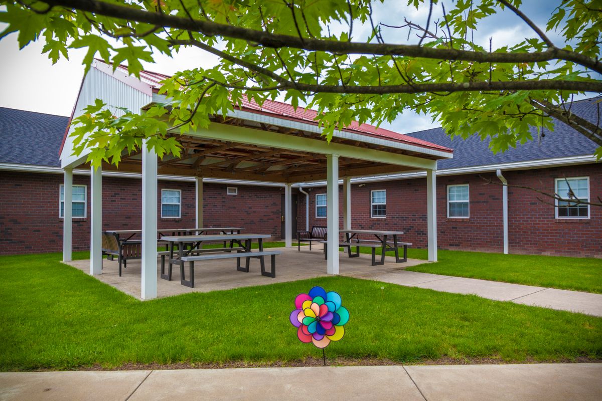 A small pavilion with picnic tables surrounded by a grassy area and trees, adjacent to a brick building.