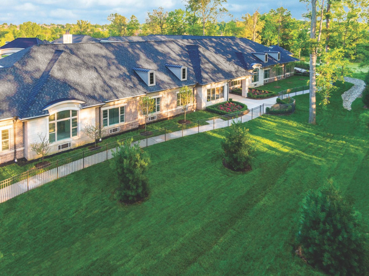 A long, elegant house with a sloped roof surrounded by a well-manicured lawn and trees.