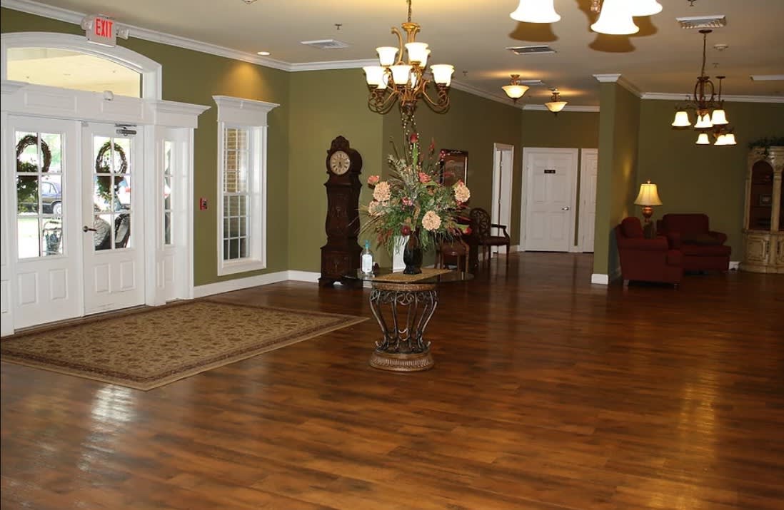 A traditional lobby with a grandfather clock, chandelier, and decorative flowers on a hardwood floor.