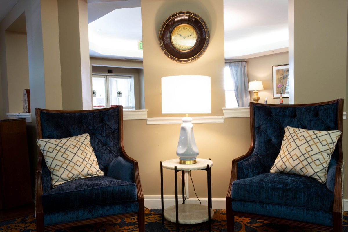 A cozy living room setup featuring two blue armchairs, a small round table with a lamp, and a wall mirror above.