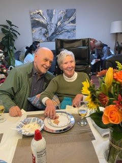 An elderly couple smiling at a table with cake and flowers, celebrating together indoors.
