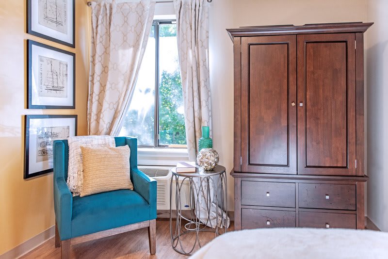 A cozy bedroom corner with a blue armchair, wooden wardrobe, and a window with light curtains.