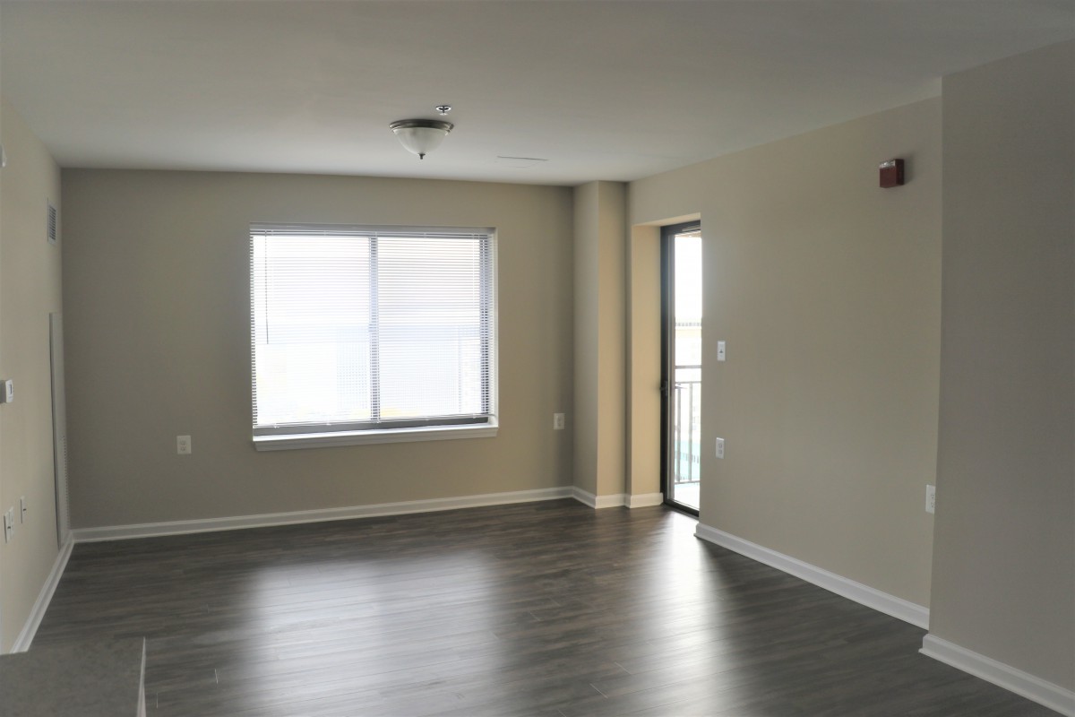 An empty apartment room with hardwood flooring, a window, and a door leading outside.