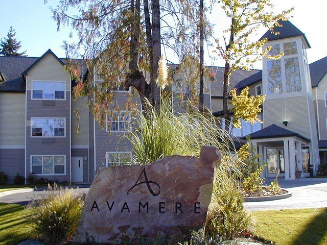 A view of a residential housing complex with a prominent stone sign in the foreground, surrounded by trees and sunlight.