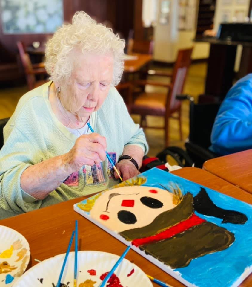 An elderly woman engaged in painting a colorful artwork at a table.
